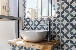 A bathroom with a white vessel sink, a chrome faucet, and a mirror above it. The wall has a geometric tile pattern. - Yellow Studio 106 | Love in Colours Collection by PikHost (Durrës)