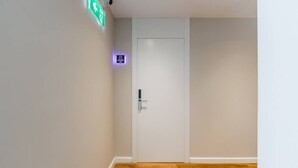 A hallway with a white door, a green emergency exit sign, and a purple sign indicating a 100% occupancy rate. - Yellow Studio 106 | Love in Colours Collection by PikHost (Durrës)