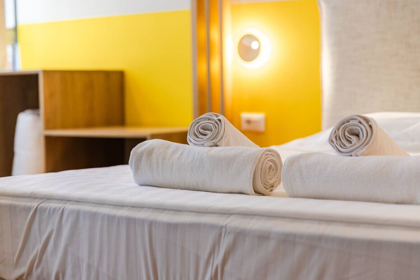 Ein Hotelzimmer mit einem Bett, zwei zusammengerollten Handtüchern, einem Holz-Nachttisch und einer Wandleuchte.