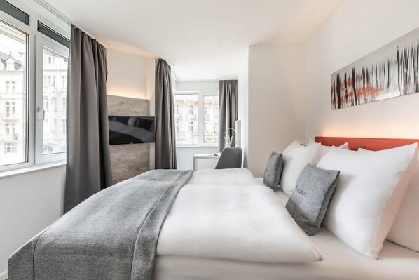 A modern hotel room with a large bed, a desk, and a view of buildings through the window. - Select Hotel Wiesbaden City (Wiesbaden)