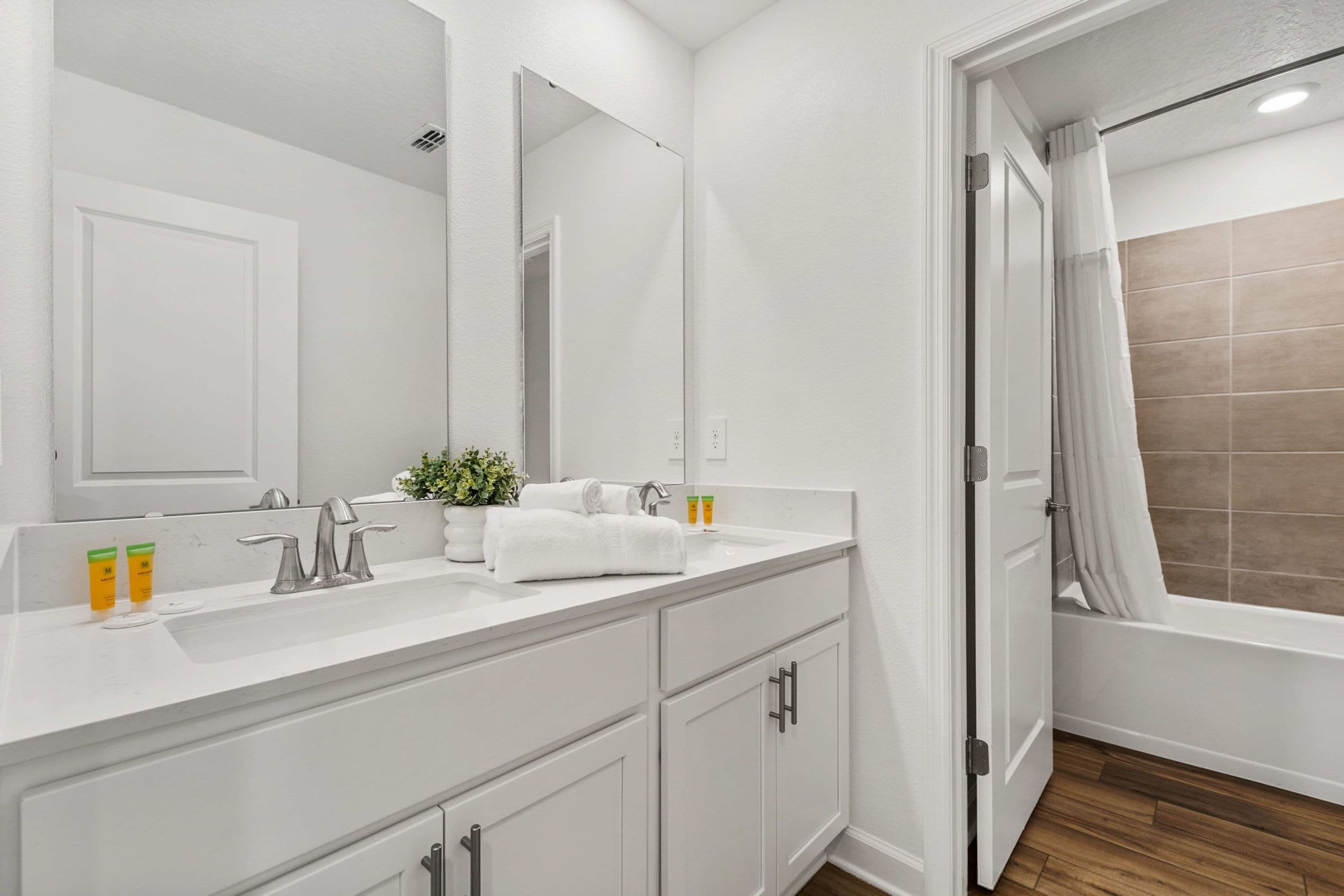 A bathroom with a white vanity, a large mirror, a sink, a shower, and a door leading to another room.
