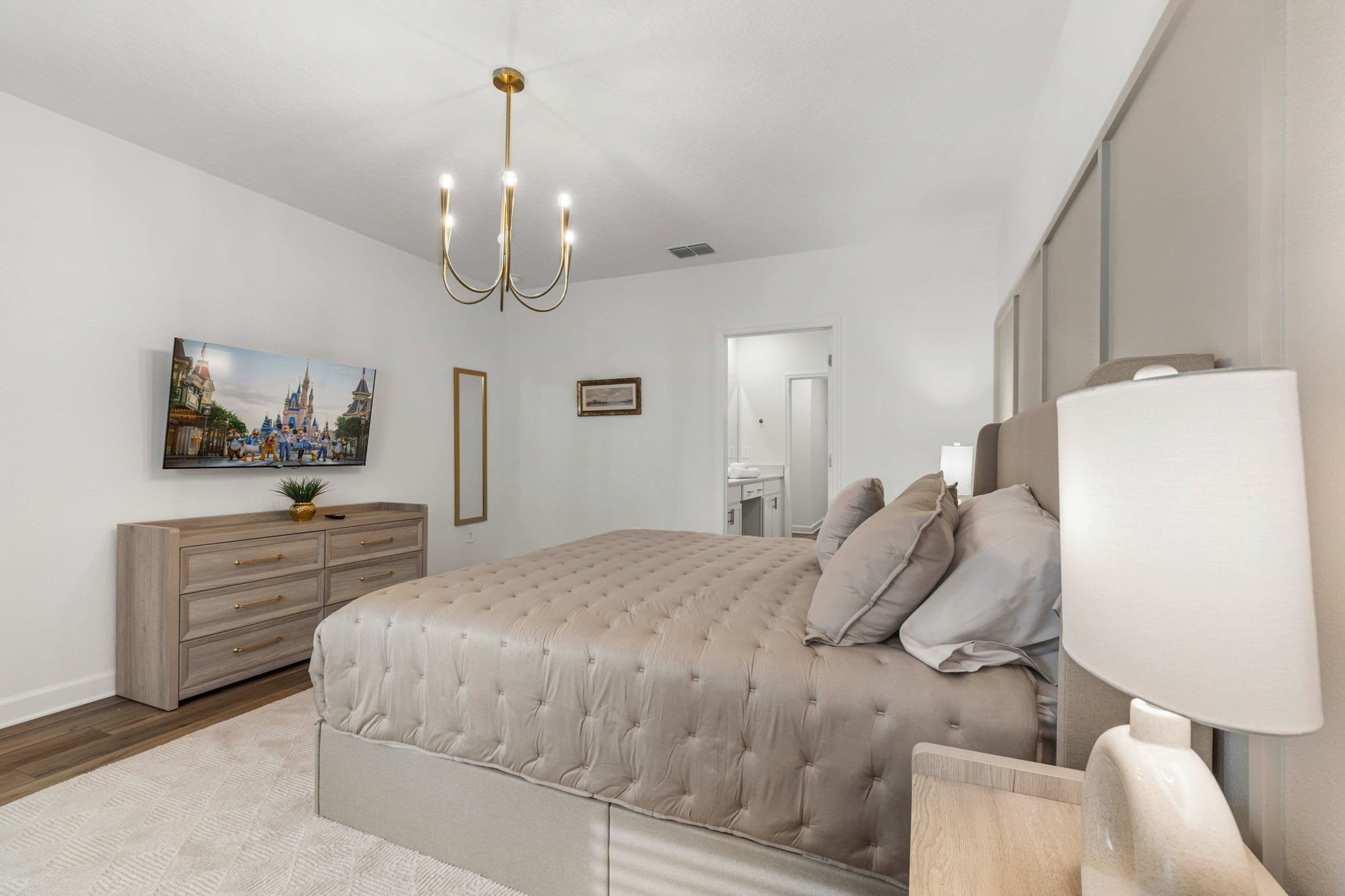 A bedroom with a tufted bed, a chandelier, a dresser, a mirror, and a painting of a castle.