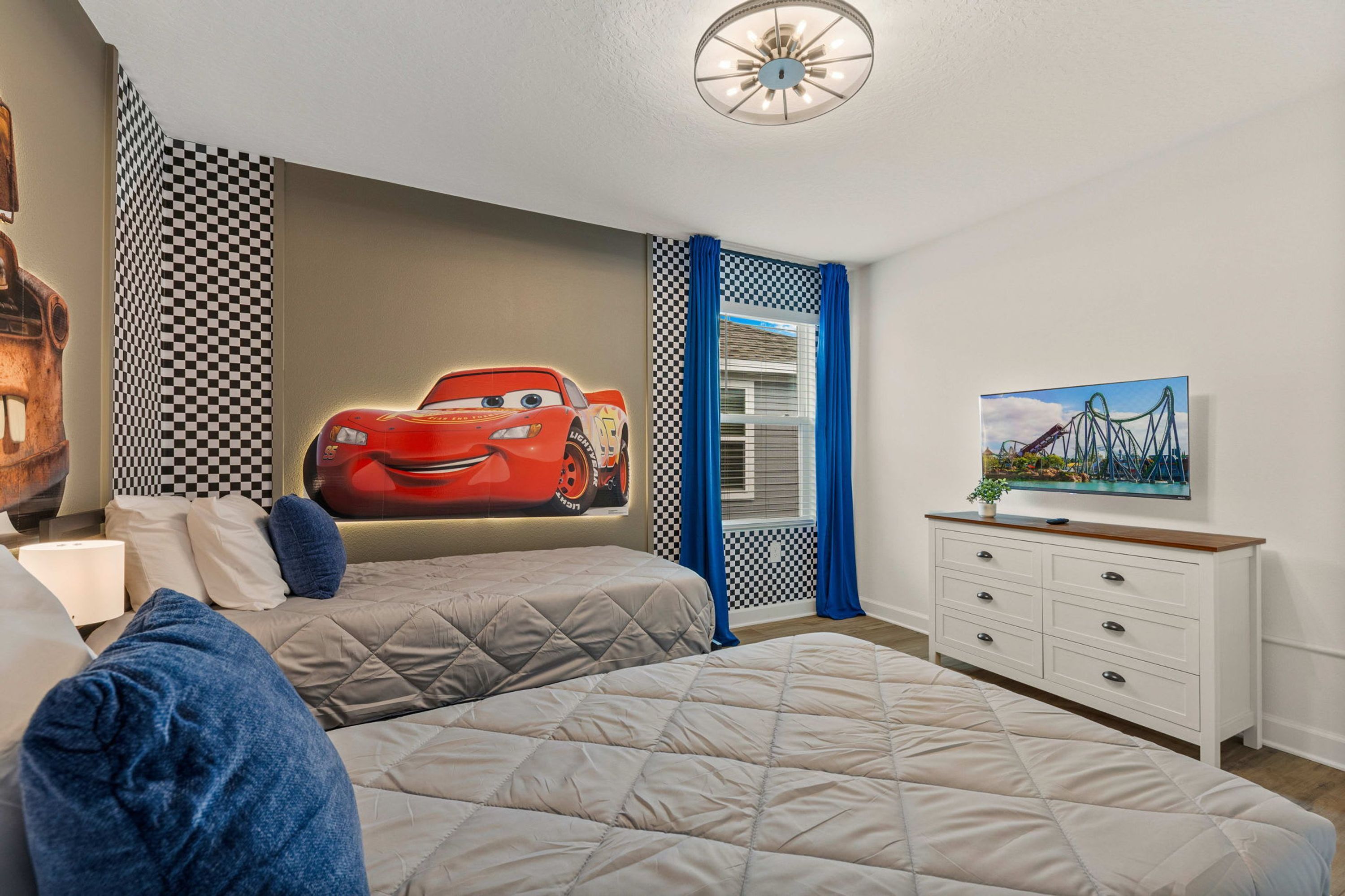 Een slaapkamer met een groot bed, een kaptafel, muurschildering van een rode auto en een raam met geruit gordijn.