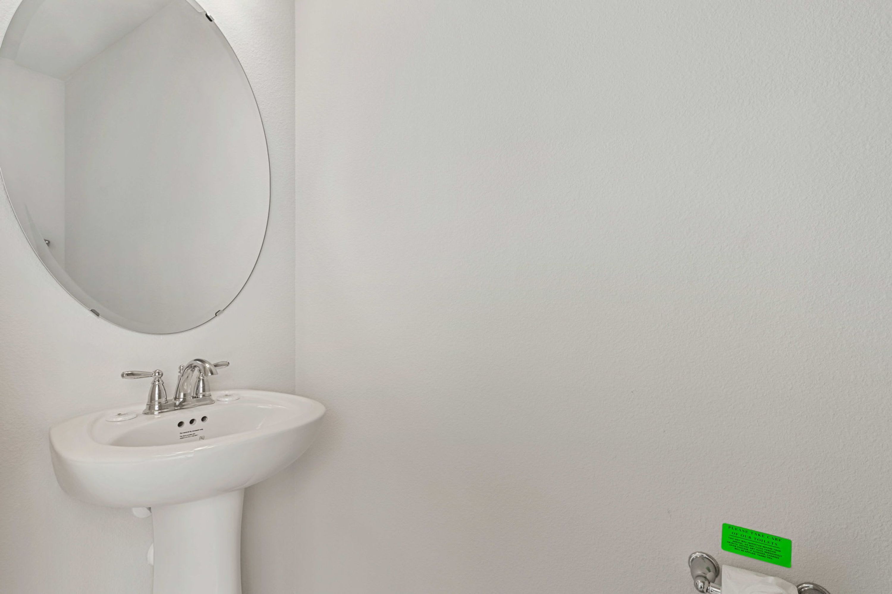 A white bathroom with a round mirror, a pedestal sink, and a green sign on the wall.