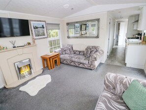 A living room with a sofa, a fireplace, a TV, and a kitchen in the background. - Meadows Farm Lodge (Swansea)