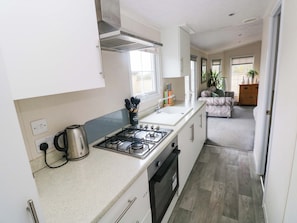 A compact kitchen with a gas stove, a kettle, and a sink. There is a window providing natural light, and a living area with a sofa and a plant.