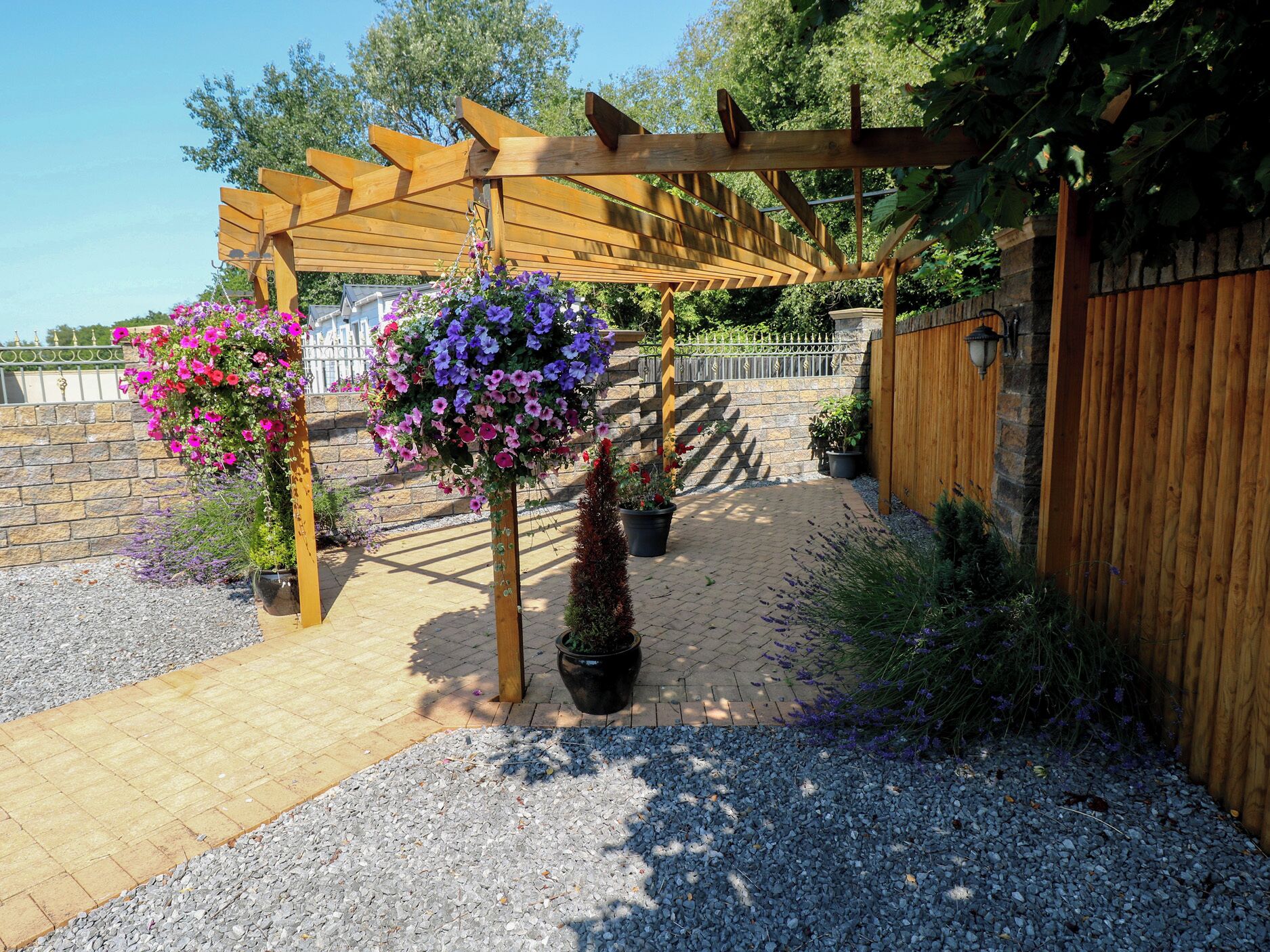 A garden with a wooden pergola, gravel pathway, and hanging flower baskets.