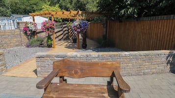 En träbänk i en trädgård, med en stenmur och en träpergola som har hängande blomkorgar.