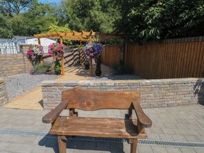 A wooden bench in a garden area with a stone wall and a wooden pergola with hanging flower baskets. - Meadows Farm Lodge (Swansea)