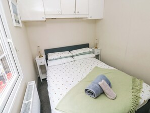 A small, neatly arranged bedroom with a bed, two bedside tables, a window, and a radiator. - Meadows Farm Lodge (Swansea)