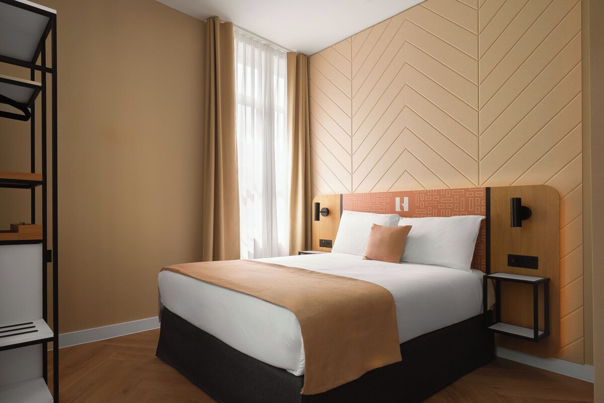 Een moderne hotelkamer met een bed, nachtkastje, houten hoofdbord, kledingkast en een raam met gordijnen.