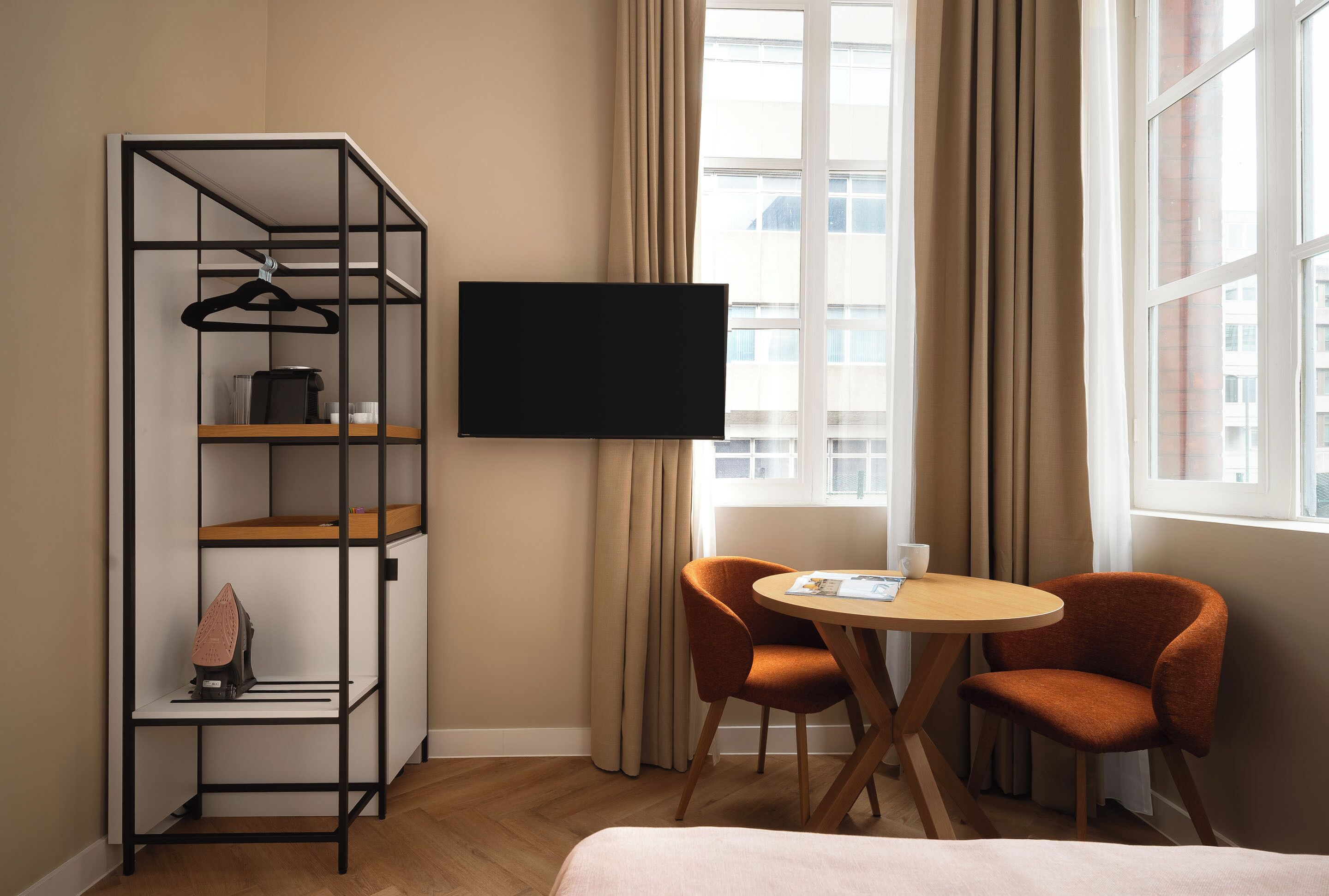 A hotel room with a TV, a wooden table, two orange chairs, a bed, and a wardrobe with a hanging rack.