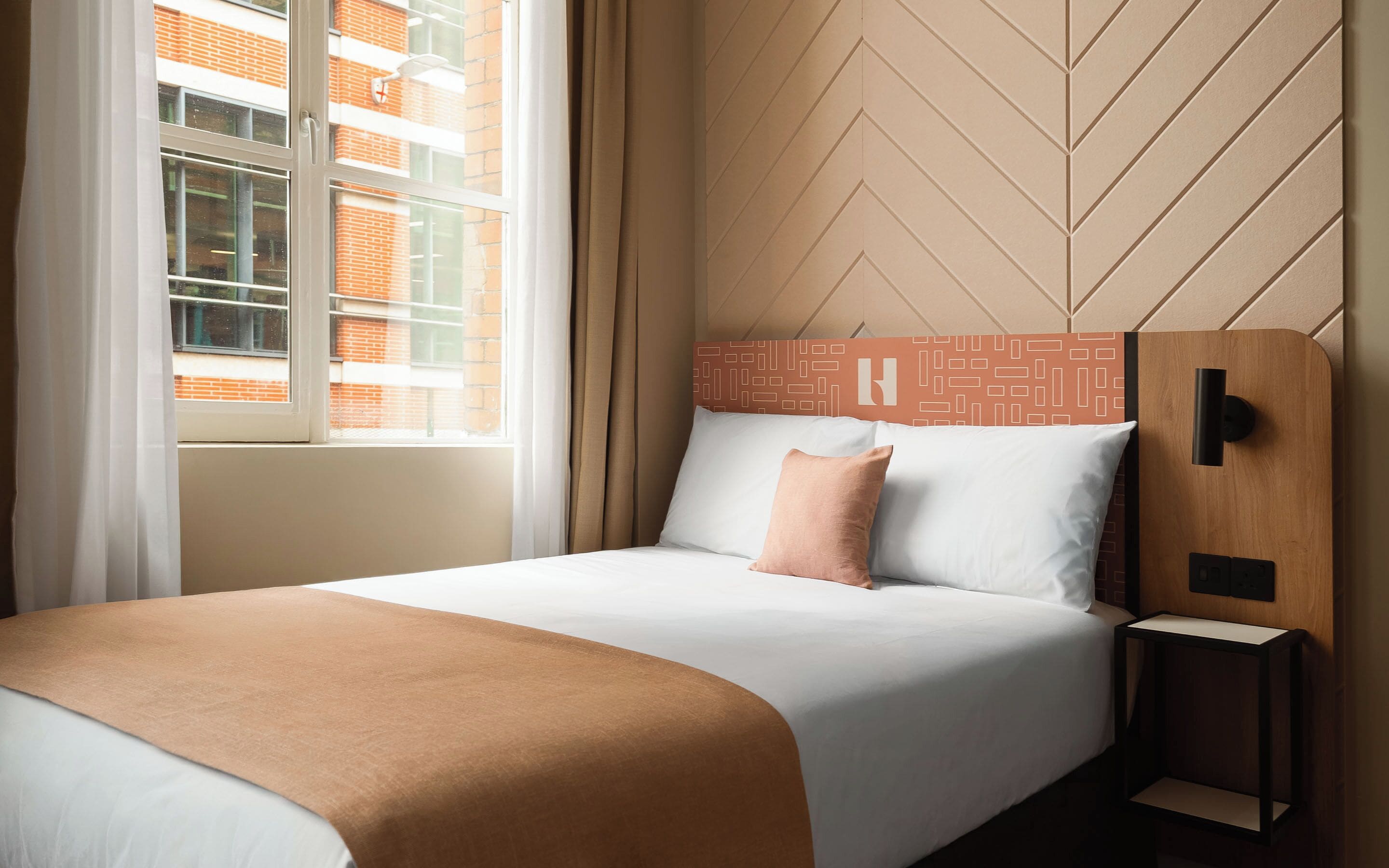 Ein Hotelzimmer mit Bett, Nachttisch, Fenster und Blick auf ein Backsteingebäude.