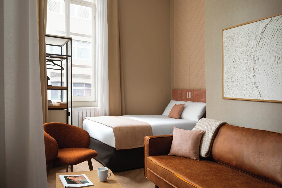 Een hotelkamer met een bed, een slaapbank, een stoel, een tafel en een raam met gordijnen.