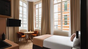 Une chambre d’hôtel avec un lit, une télévision, une petite table avec des chaises et de grandes fenêtres avec des rideaux.