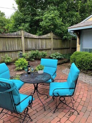 Eine Terrasse mit einem runden Tisch und vier blauen Stühlen, umgeben von einem Holzzaun und viel Grün.