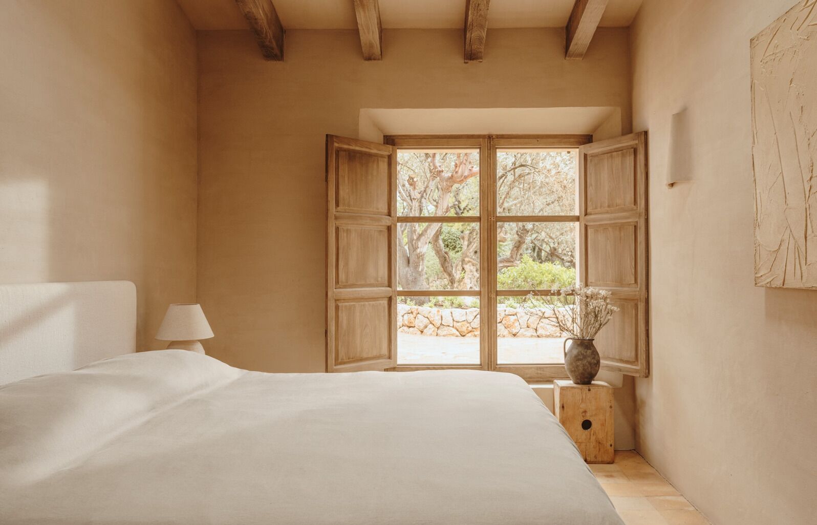 A bedroom with a large bed, a window with wooden shutters, a bedside table with a lamp, and a vase with flowers.