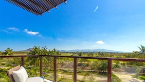 A balcony with a wicker chair, a glass railing, and a view of a green landscape with palm trees and mountains. - Condo Ara in Punta Mita (Punta de Mita)