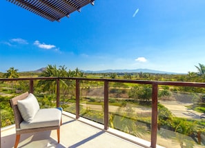 A balcony with a wicker chair, a glass railing, and a view of a green landscape with palm trees and mountains. - Condo Ara in Punta Mita (Punta de Mita)