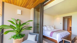 Ein modernes Hotelzimmer mit einem Bett, einer Sitzecke inklusive Sessel und Tisch, einer Topfpflanze und einer Schiebeglastür, die auf einen Balkon führt.
