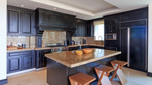 A modern kitchen with dark cabinetry, granite countertops, a central island with stools, and built-in appliances. - Condo Aralia in Punta Mita (Punta de Mita)