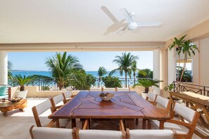 A wooden dining table with white cushioned chairs, a ceiling fan, and a view of the ocean and palm trees. - Condo Peonia in Punta Mita (Punta de Mita)