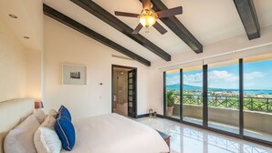Ein Schlafzimmer mit einem großen Bett, einem Deckenventilator, Blick durch das Fenster und einer Tür zu einem Badezimmer.