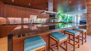 Une cuisine moderne dotée d’un îlot central, de tabourets de bar en bois et offrant une vue sur la verdure grâce à un mur de verre.