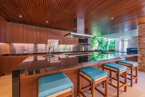 A modern kitchen with a central island, wooden bar stools, and a view of greenery through a glass wall. - Estate Taurus in Kupuri Oceanfront Private Pool Design (Punta de Mita)