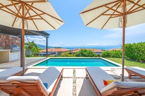 A poolside area with lounge chairs and umbrellas, a barbecue grill, and a view of the ocean and buildings. - Villa Perla in Punta Mita (Punta de Mita)