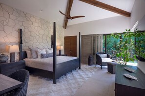 A bedroom with a stone wall, a four-poster bed, a ceiling fan, a large window with a view of greenery, and a wicker chair. - Villa Tamayo in Punta Mita (Punta de Mita)