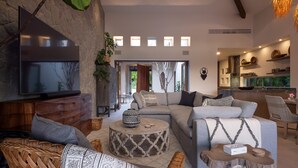 Ein modernes Wohnzimmer mit Fernseher, grauer Couch, Korbmöbeln und einer Küche im Hintergrund.