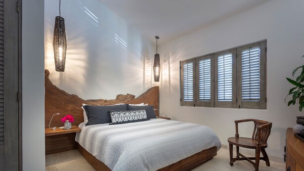 Ein Schlafzimmer mit einem hölzernen Kopfteil, einem Bett mit weißer Decke, zwei Nachttischlampen, einem Nachttisch aus Holz mit einem Blumenstrauß und einem Fenster mit Jalousien.