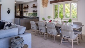 Ein modernes Wohnzimmer mit einem grauen Sofa, einem Esstisch mit Korbstühlen, einer Küche im Hintergrund und einem großen Fenster mit Blick auf Grünflächen.