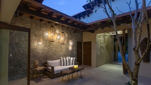 Eine Steinsäule, eine Holzbank, ein Sofa, hängende Lampen und ein Baum in einem Innenhof.