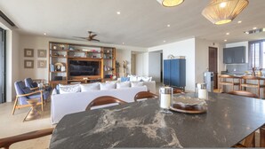 A spacious living area with a large black marble table, a white sofa, and a television. There are wooden shelves with various items, a ceiling fan, and a kitchen area visible. - Condo Kumar in Punta Mita (Punta de Mita)
