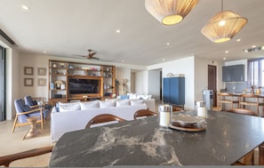 Ein geräumiger Wohnbereich mit einem großen schwarzen Marmortisch, einem weißen Sofa und einem Fernseher. Holzregale mit diversen Gegenständen, ein Deckenventilator und ein sichtbarer Küchenbereich sind ebenfalls vorhanden.