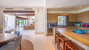 A modern kitchen with wooden cabinets, a central island with stools, and a dining area with a round table and chairs. - Casa Marlago in Punta Mita (Punta de Mita)