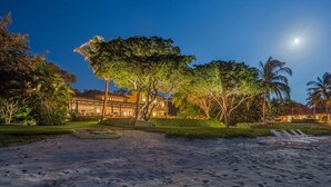 Una propiedad frente a la playa con un edificio bien iluminado, palmeras y una zona de arena con sillas de descanso.