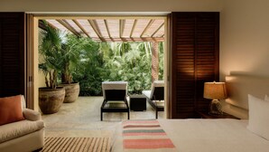 Une chambre à coucher comprenant un lit, deux chaises, un canapé, une lampe et une vue sur un jardin luxuriant, visible à travers une porte ouverte.
