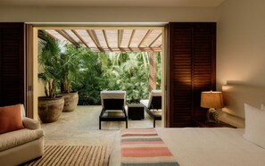 A bedroom with a bed, two chairs, a sofa, a lamp, and a view of a lush garden through an open door. - Estate Jaguar in Punta Mita (Punta de Mita)
