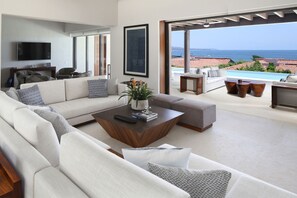 Ein modernes Wohnzimmer mit einer großen Couch, einem Couchtisch und einem Fernseher. Durch große Fenster ist der Meerblick zu sehen.