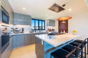 A modern kitchen with grey cabinets, a large island, and a view of a house through the windows. - Condo Sardan in Punta Mita (Punta de Mita)