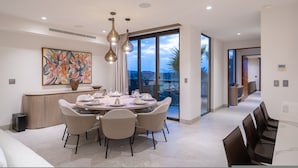A modern dining area with a round table, chairs, pendant lighting, a large painting, and a view through glass doors. - Condo La Joya in Punta Mita (Punta de Mita)