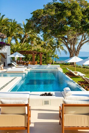 Ein Swimmingpool mit Liegestühlen und Sonnenschirmen, umgeben von Palmen und Blick aufs Meer.