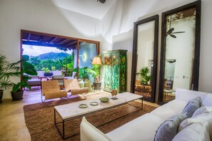 A living room with a sofa, chairs, a coffee table, and a view of a mountainous landscape through a glass door. - Penthouse Pacifico in Punta Mita (Punta de Mita)