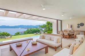 A spacious living area with a large window overlooking a pool and mountains, a dining area with a table and chairs, and a sofa set. - Estate Ikal in Punta Mita (Punta de Mita)
