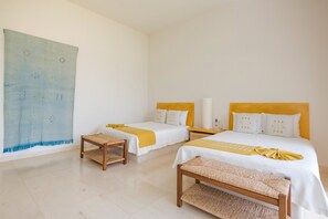 Ein Hotelzimmer mit zwei Betten, einer gewebten Bank, einem kleinen Tisch und einem großen, abstrakten Gemälde an der Wand.