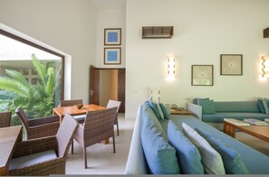 Ein Wohnzimmer mit einer Couch, einem Esstisch mit Stühlen, einem großen Fenster mit Blick auf Grünflächen und Wandleuchten.