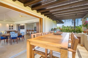 A spacious dining area with a long wooden table and blue upholstered chairs, a ceiling fan, and a view of a lush garden. - Condo Lago in Punta Mita (Punta de Mita)